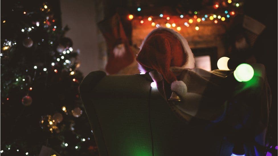 A christmas living room scene, with a Santa red bobble hat poking above the back of a sofa and a Christmas tree in the corner.